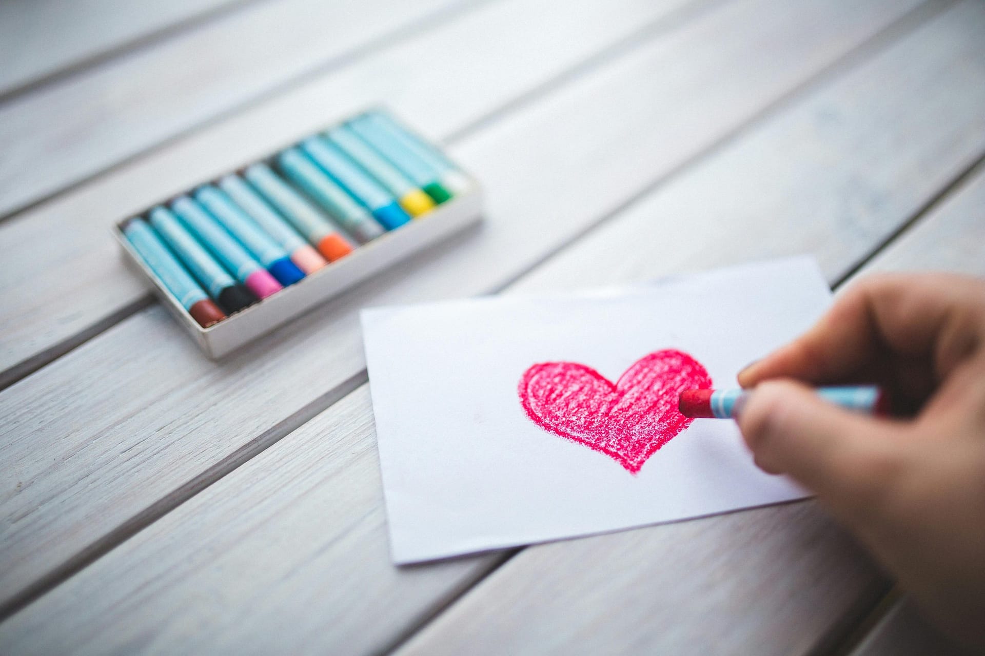 A close-up of a hand using pastel crayons to draw a red heart on paper.
