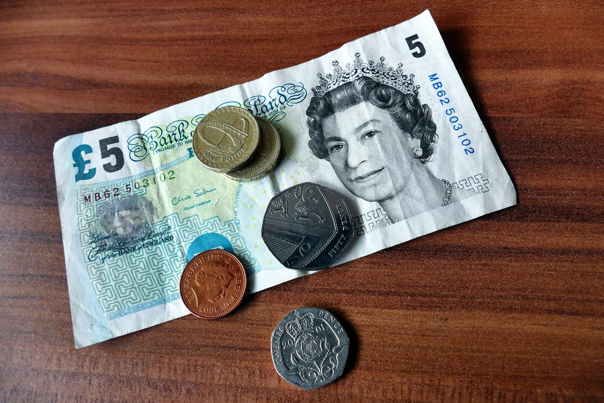 Close-up of a five-pound note and coins representing UK currency on a wooden surface.